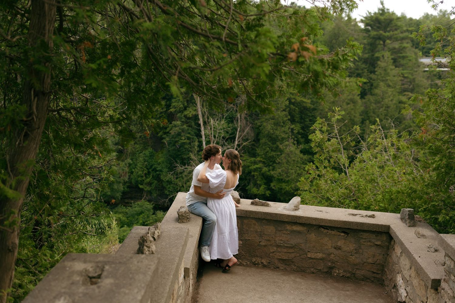 Couple sharing a romantic kiss on a stone balcony overlooking lush green forest trees.