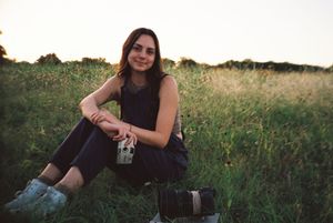 Young woman sitting in a lush green field at sunset, holding a vintage camera, wearing a black sleeveless outfit.