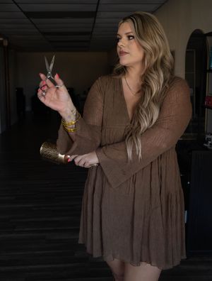 A blonde female hairstylist in a brown dress holds scissors and a brush in a salon setting.