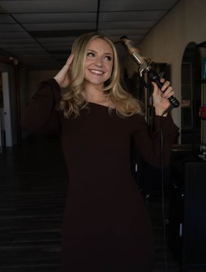 Smiling blonde woman in dark dress holding a curling iron while styling her hair in a salon hallway.