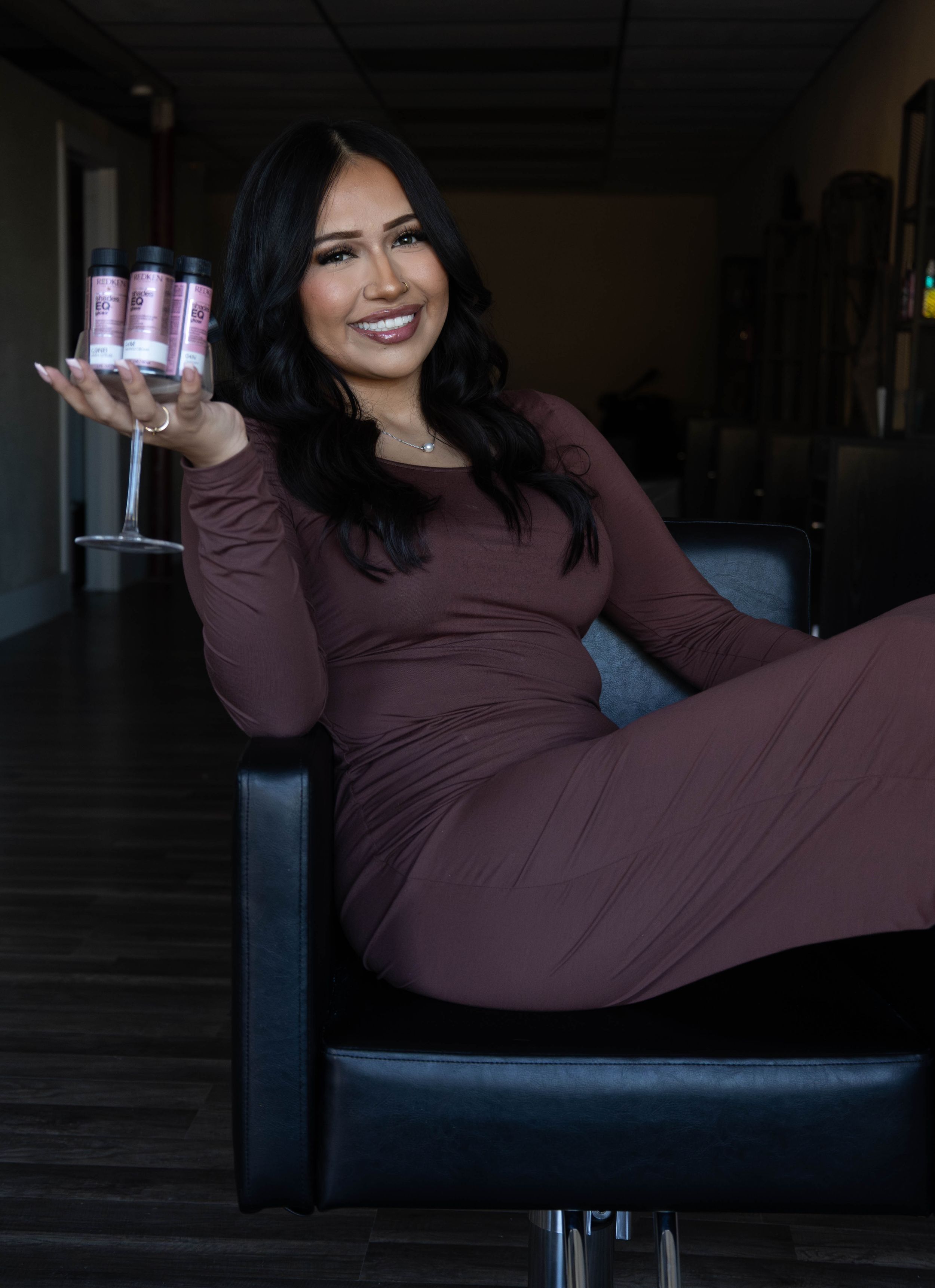 Woman in burgundy dress sitting in salon chair holding hair care products, smiling confidently.