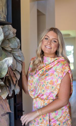Smiling blonde woman in colorful floral dress poses by a decorative leaf wreath on a dark door.
