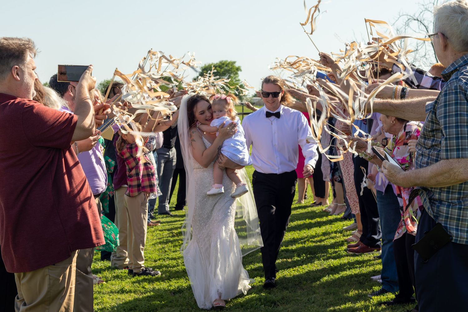 Bride and groom walk through ribbon wand sendoff with guests cheering at outdoor wedding reception.