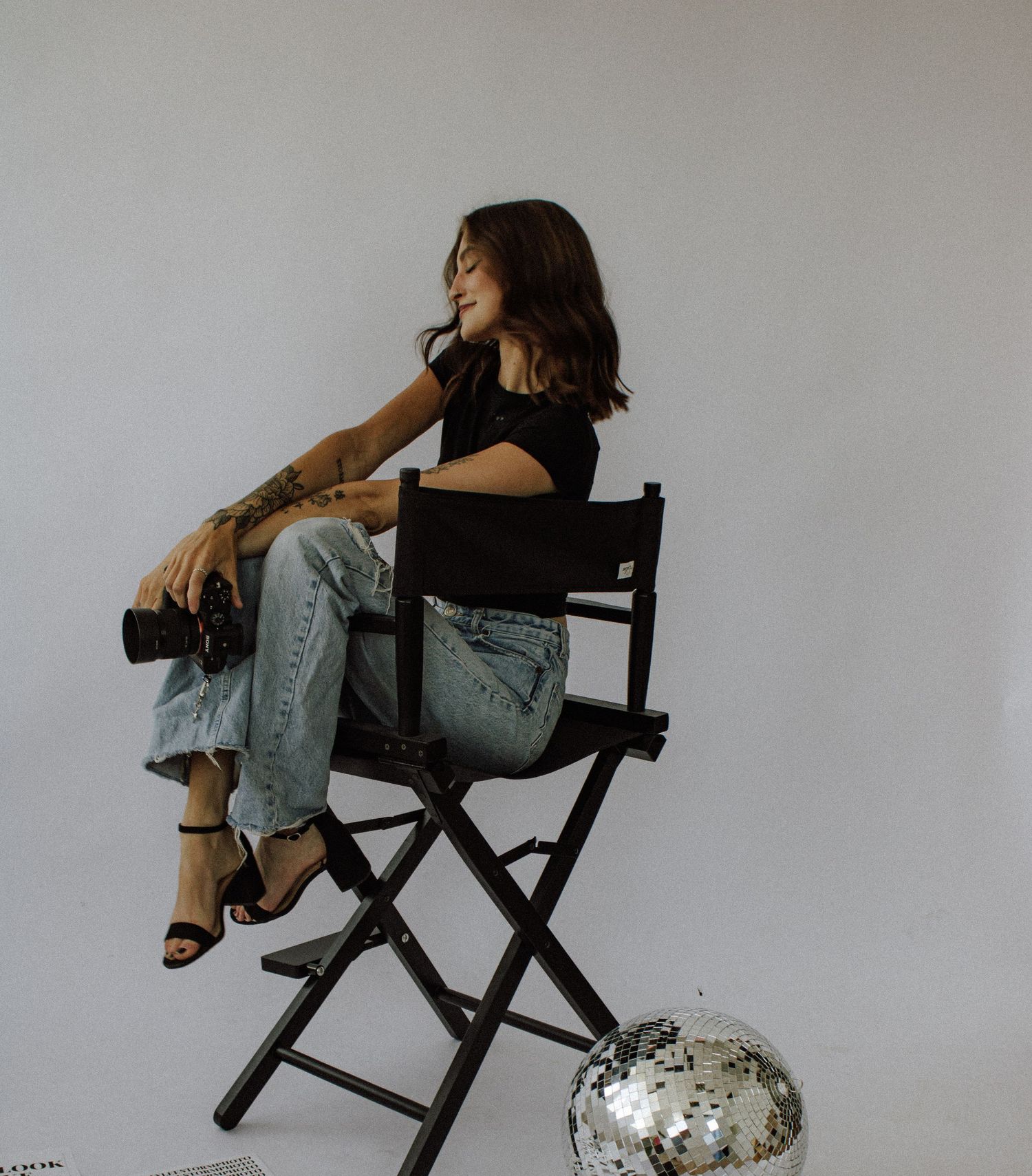 Woman in black top and jeans sitting on a director's chair next to a disco ball on white background. Kingsport, TN