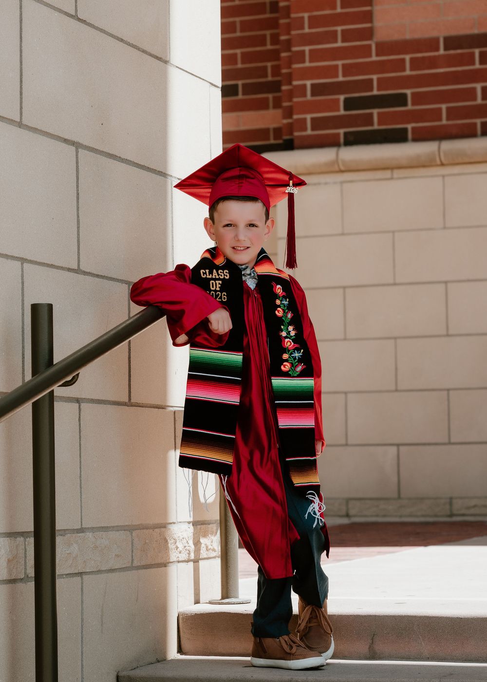Young boy in red graduation cap and gown with colorful serape stole smiling on steps at graduation ceremony.