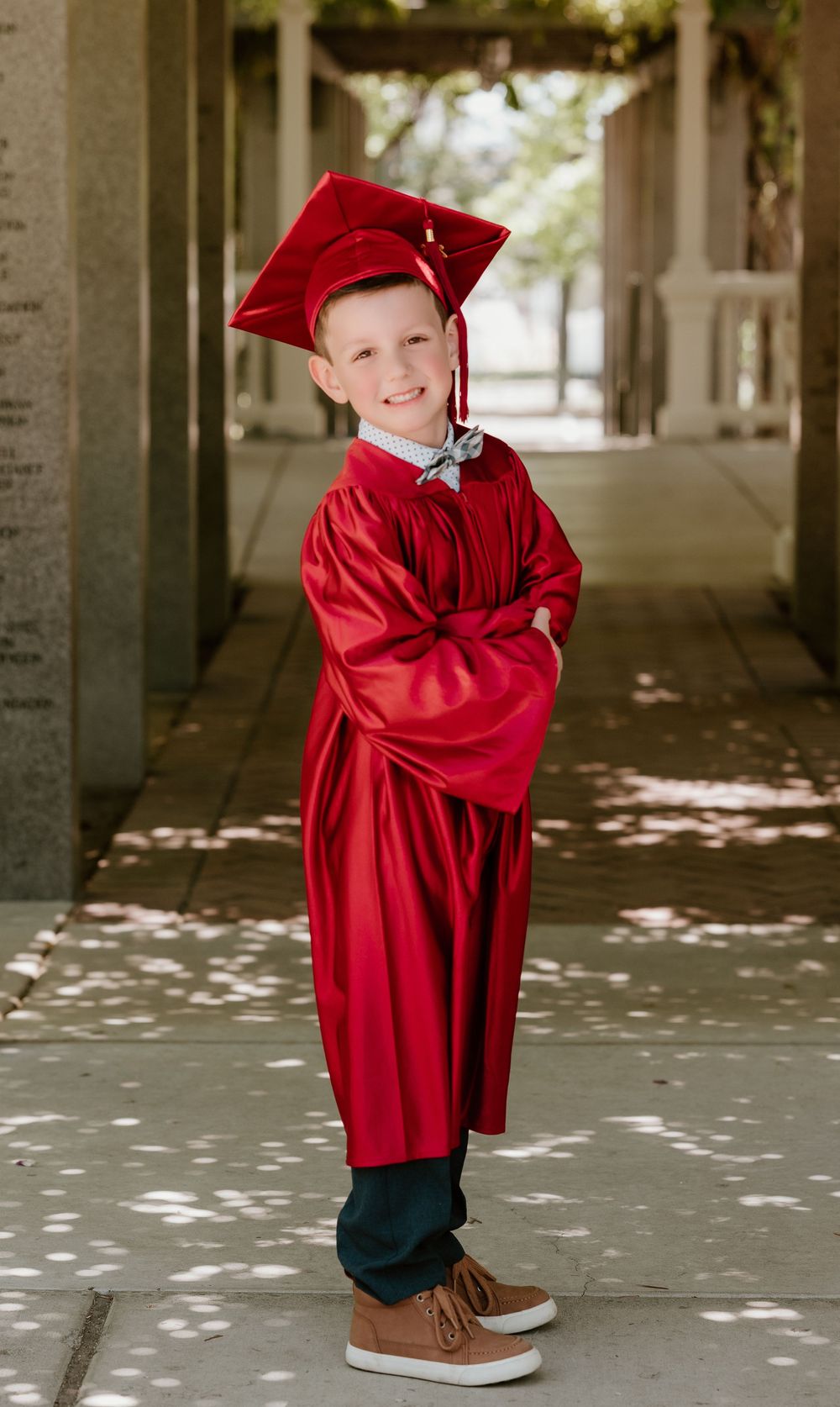 Young boy in red graduation cap and gown smiling with arms crossed outdoors.