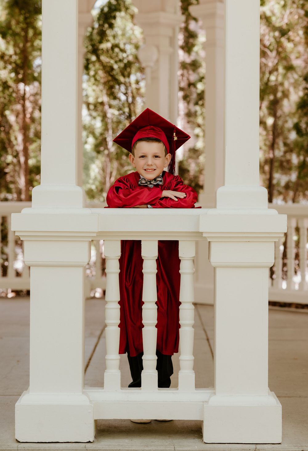 Young boy in red graduation cap and gown smiling between white columns at an outdoor ceremony.