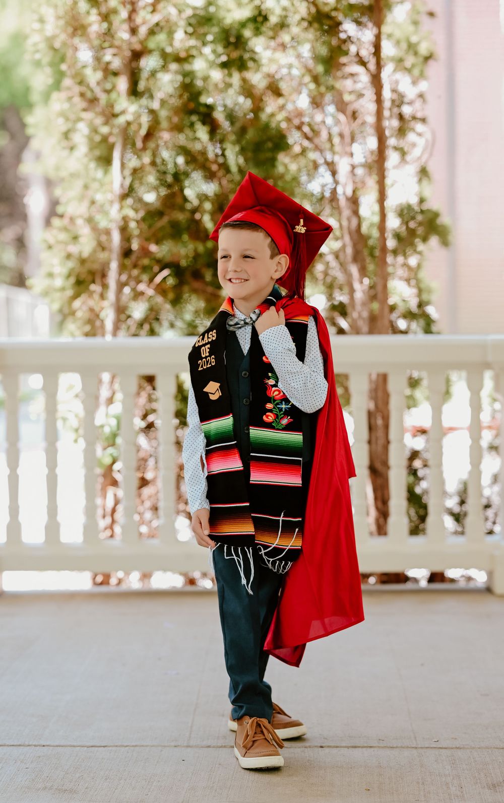 Young boy smiling in red graduation cap and gown with colorful sash, posing outdoors on a porch.