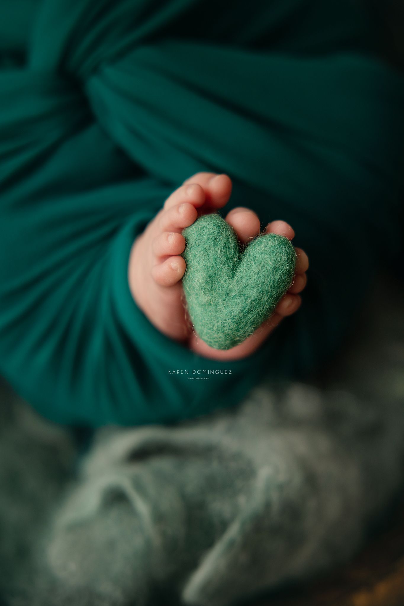 Newborn baby toes holding a small green felt heart, wrapped in teal fabric, in a soft focus photography portrait.