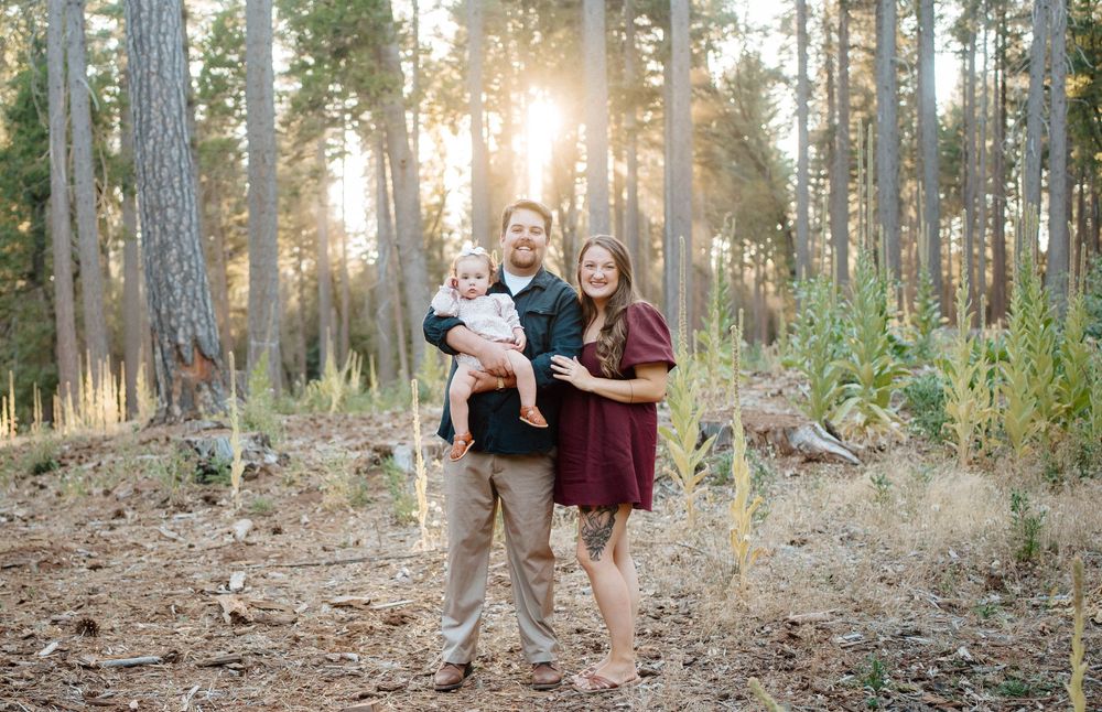 Family of three posing in sunlit pine forest, parents smiling holding baby, golden hour light filtering through tall trees.