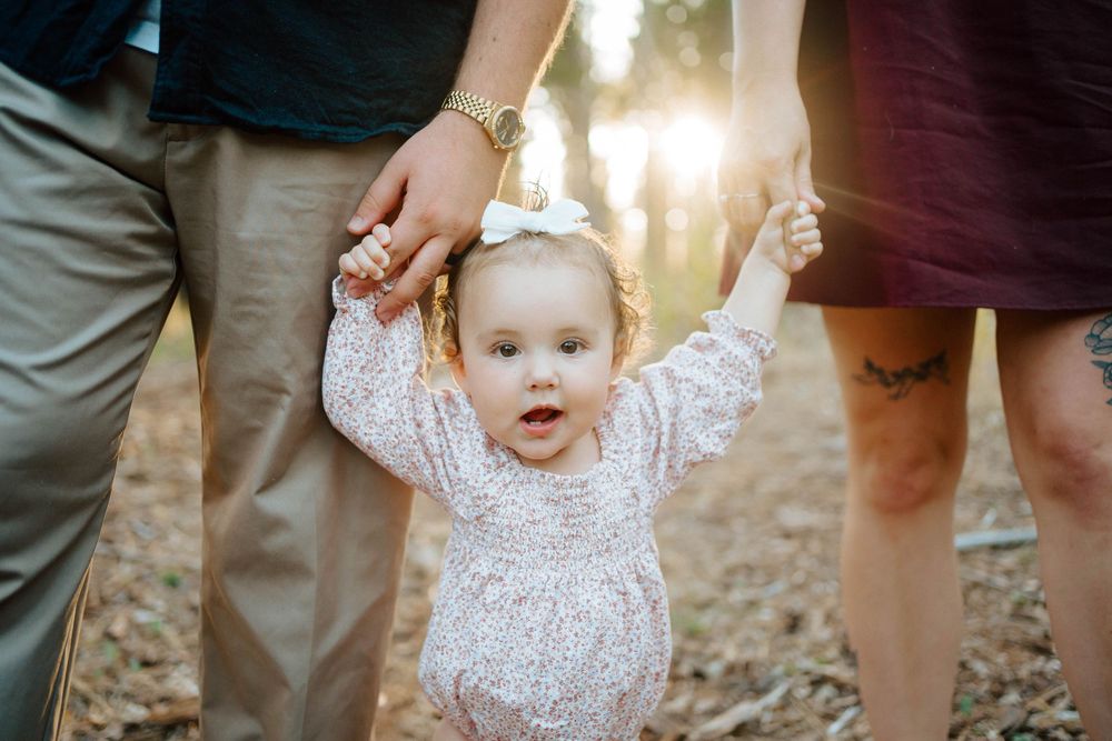 A joyful baby girl in a floral romper holds her parents hands outdoors in golden sunlight.