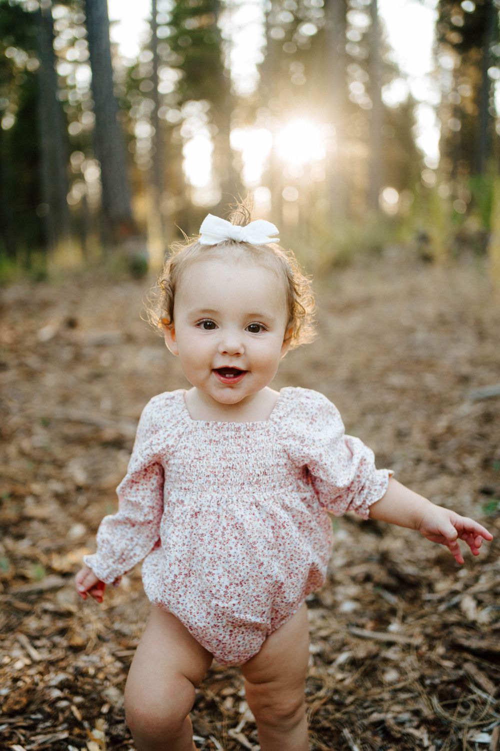 Smiling baby girl in floral romper with white bow hair clip standing outdoors in forest during golden sunset.