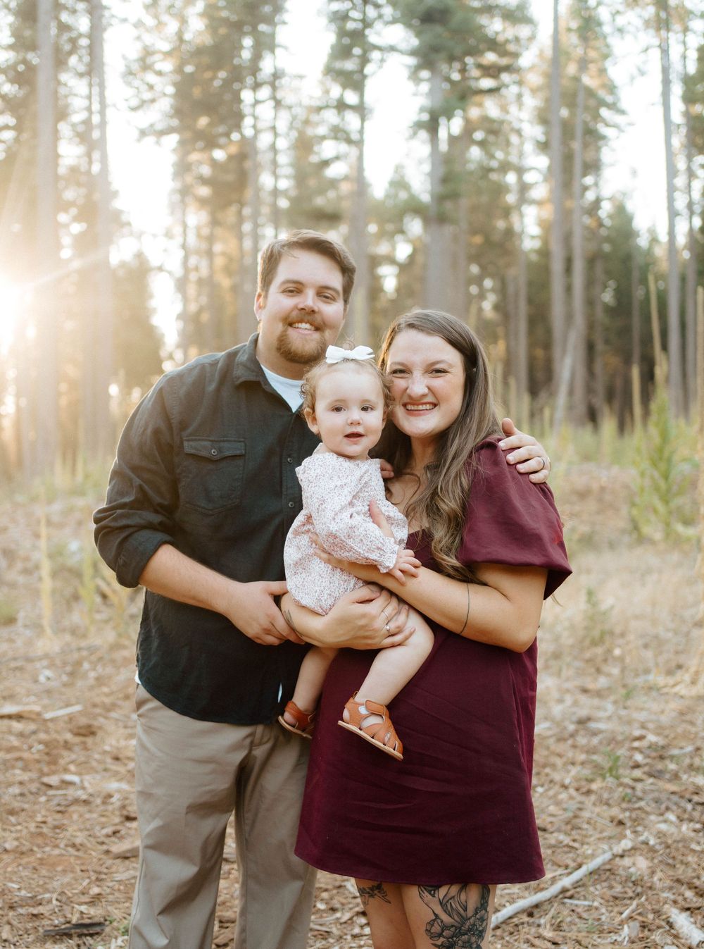 A young family poses outdoors in a sunlit forest, with parents holding their smiling baby girl in a floral dress.