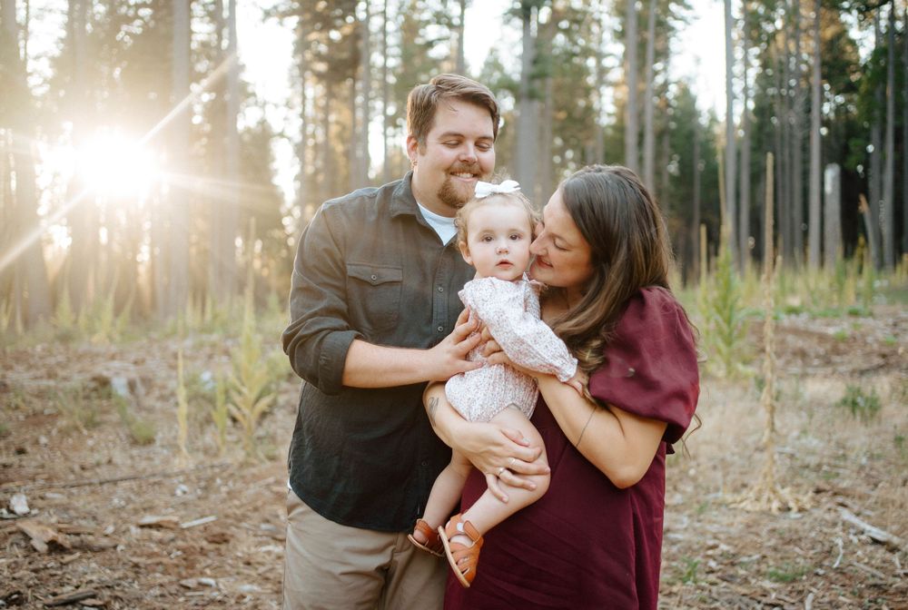 A happy family with a baby girl poses in a sunlit pine forest during a golden hour outdoor photo session.