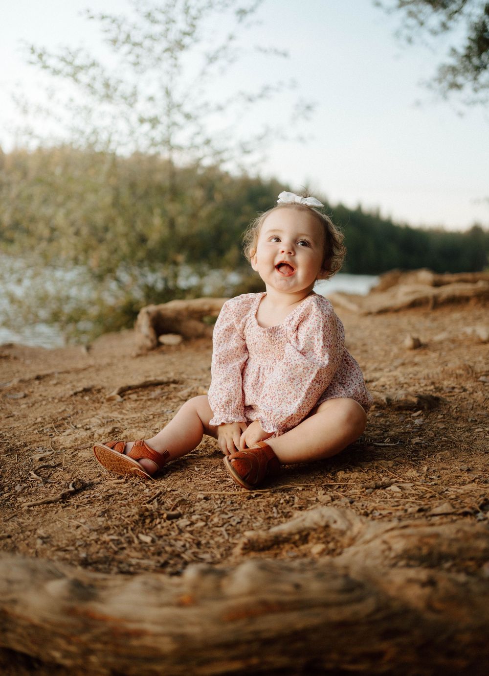 A happy baby girl in a pink floral romper and brown shoes sits on a log outdoors near a lake.