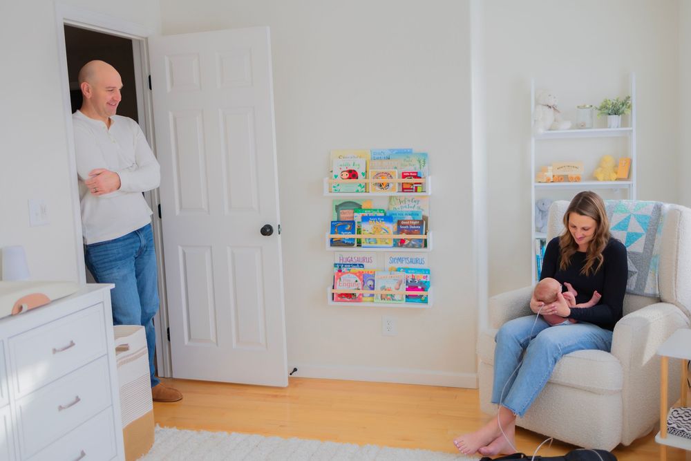 Parents in a cozy nursery, mother sitting in glider holding newborn baby while father stands in doorway smiling.