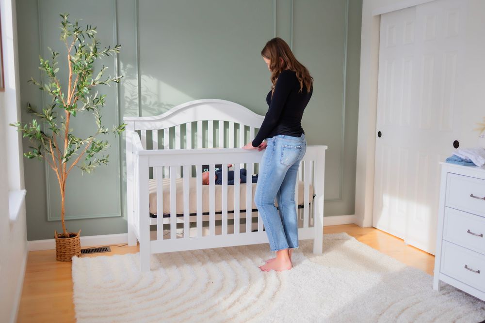 Woman in jeans arranging a white baby crib in a green nursery room with hardwood floors and a decorative tree.
