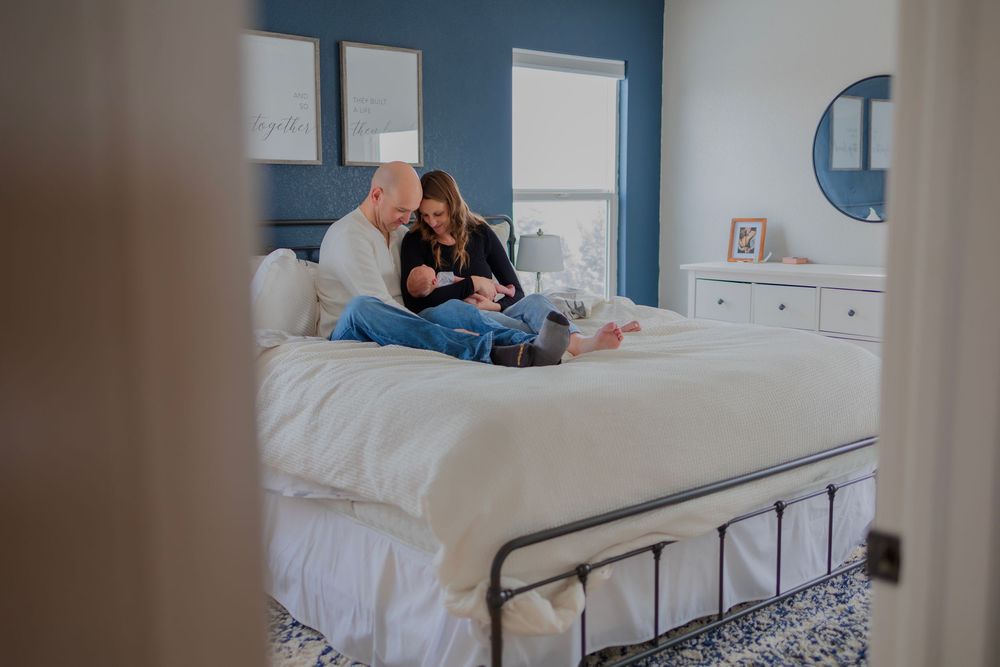New parents sit on a white bed in a blue bedroom, lovingly cradling their newborn baby together.