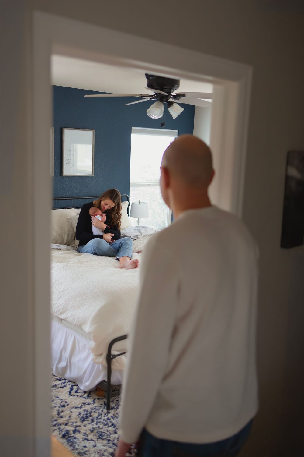 A man stands in a doorway watching a woman cuddle a baby on a white bed in a blue bedroom.