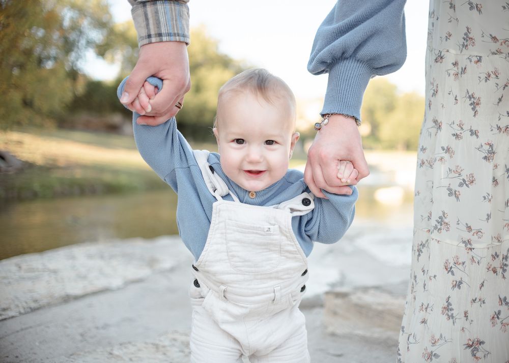 Baby boy learning to walk while holding parents hands at Nolan Creek in Temple Texas during family photoshoot