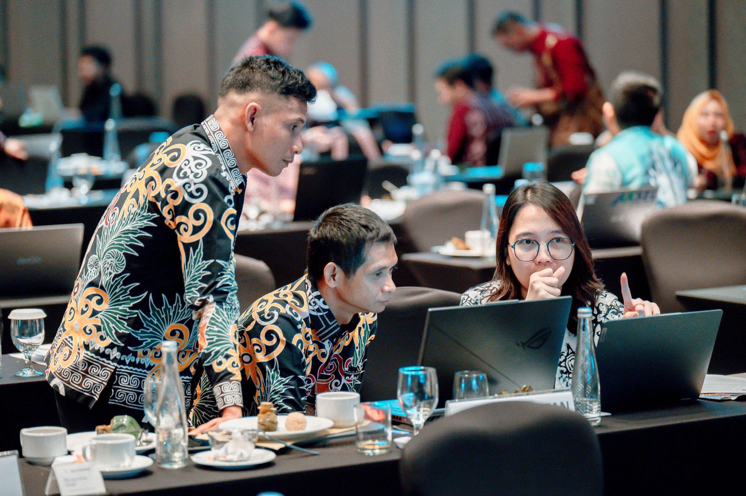Professionals in batik attire collaborate at a conference table with laptops in a modern seminar room.