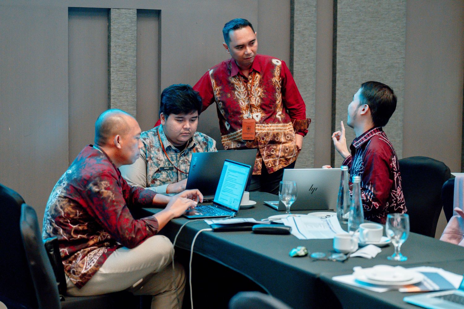 Four professionals in batik shirts collaborate around a table with laptops during a business meeting.