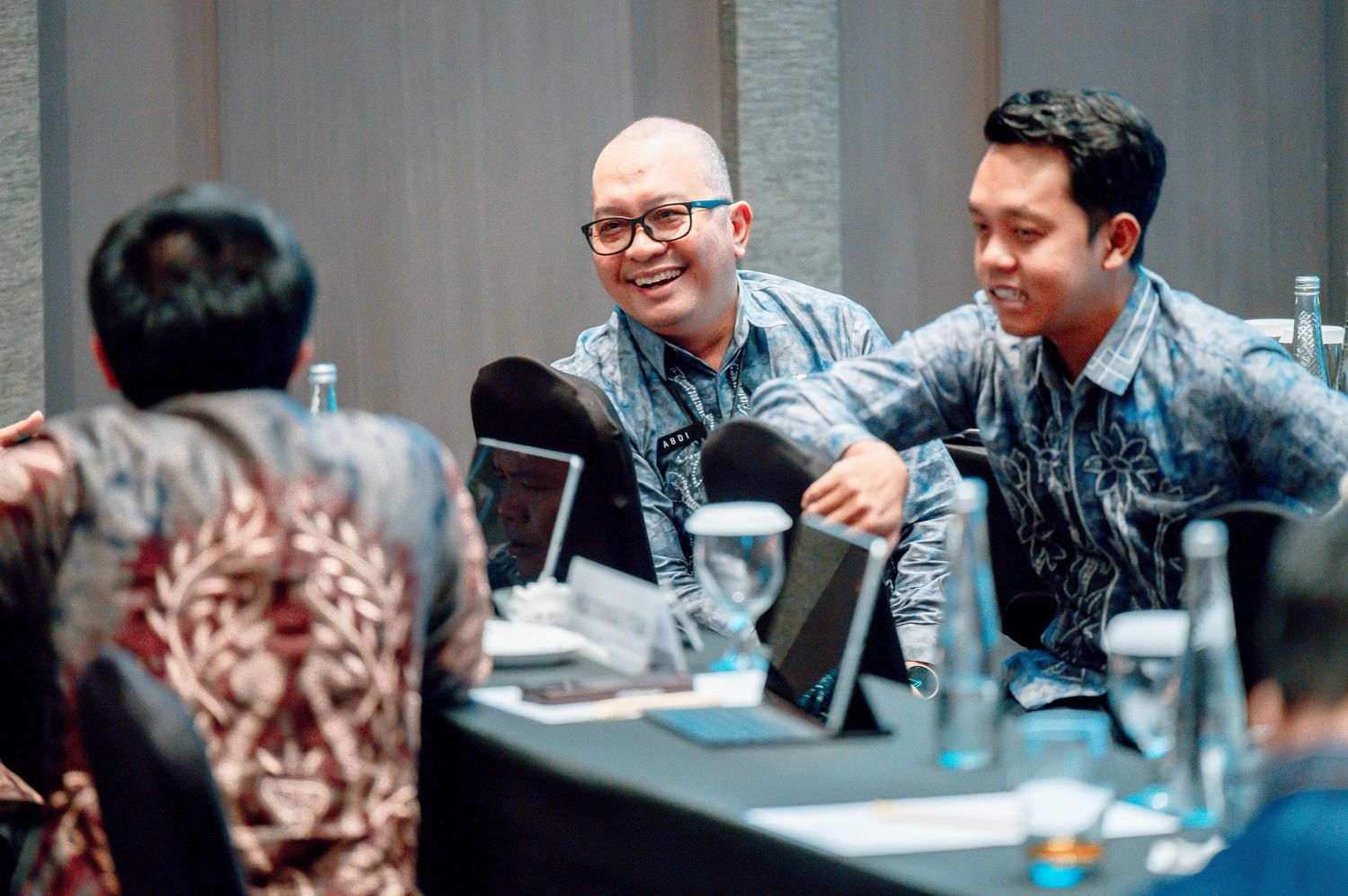 Two men in batik shirts smile during a professional meeting at a conference table with laptops and water bottles.