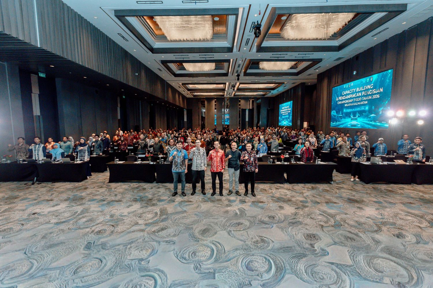 Large corporate conference gathering with hundreds of attendees in a modern ballroom venue with blue stage lighting.