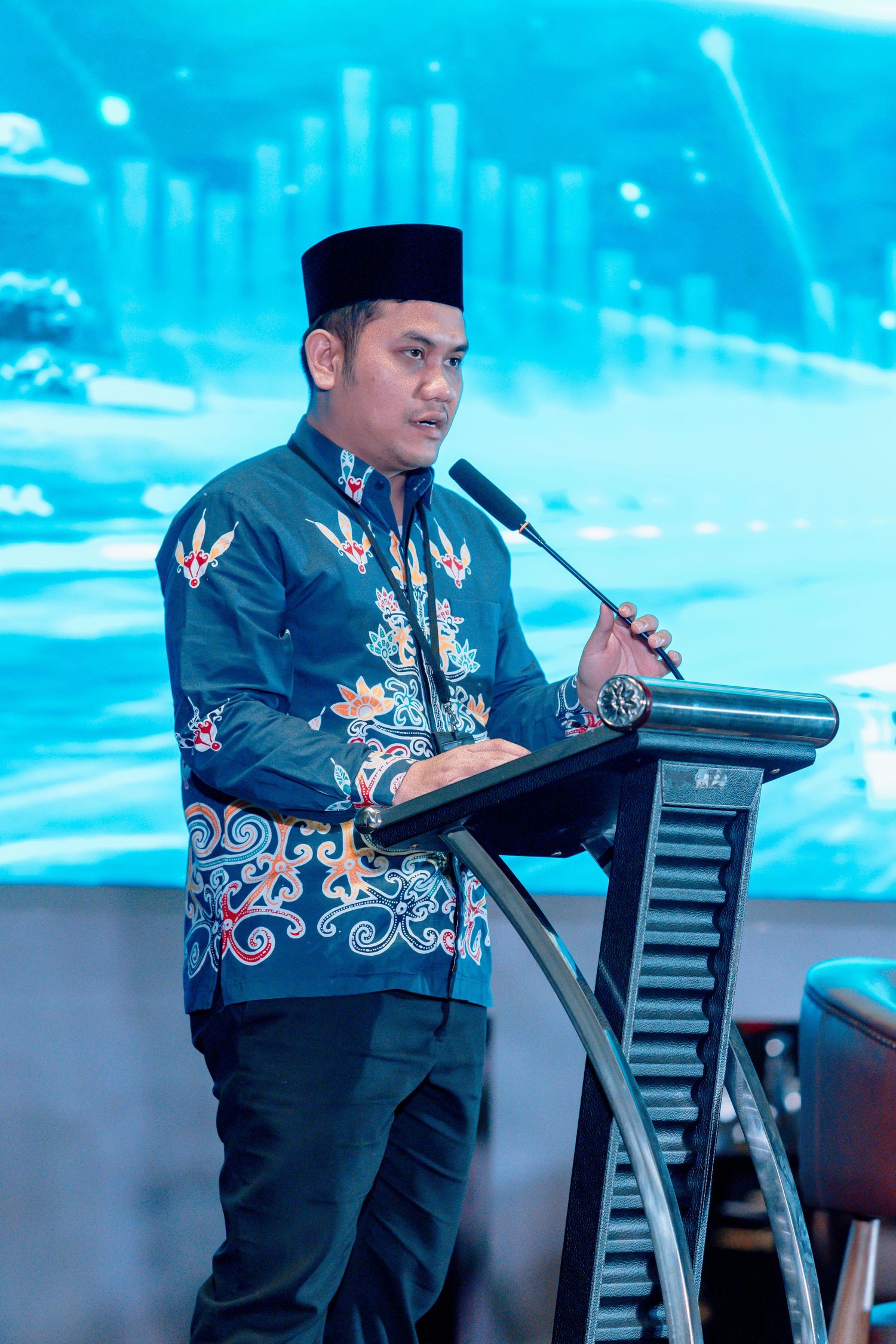 Man in batik shirt and black cap speaking at podium with microphone at formal event.