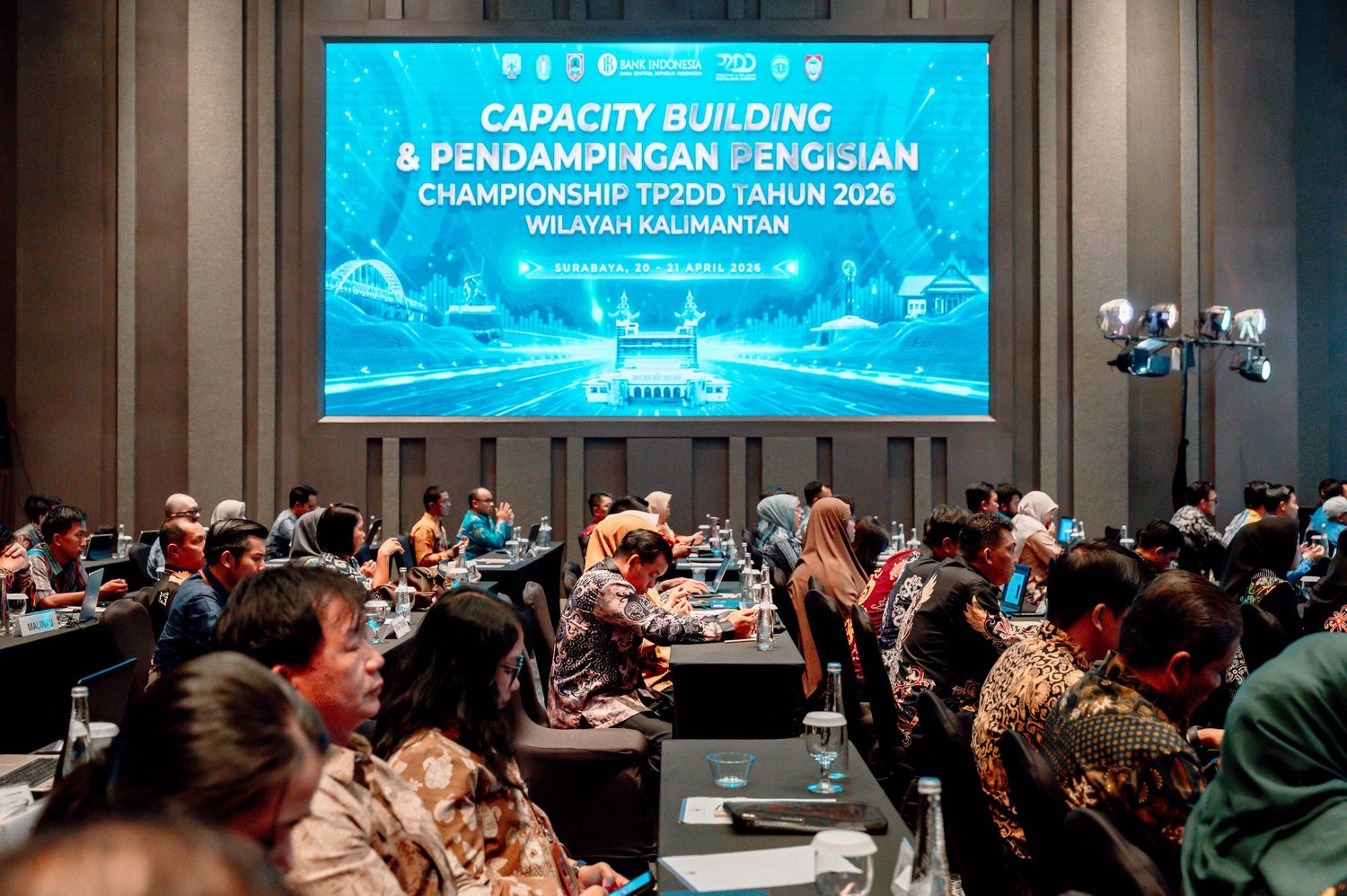 Capacity building seminar in Kalimantan with attendees seated at tables facing a large LED screen displaying event details.