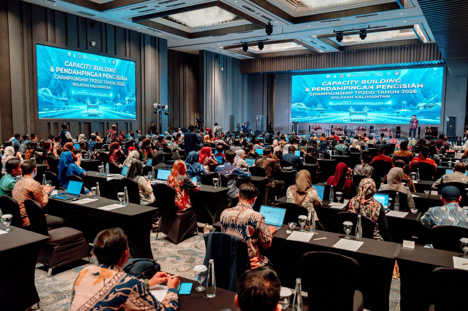 Large conference hall filled with attendees at a professional seminar with blue presentation screens displayed on stage.