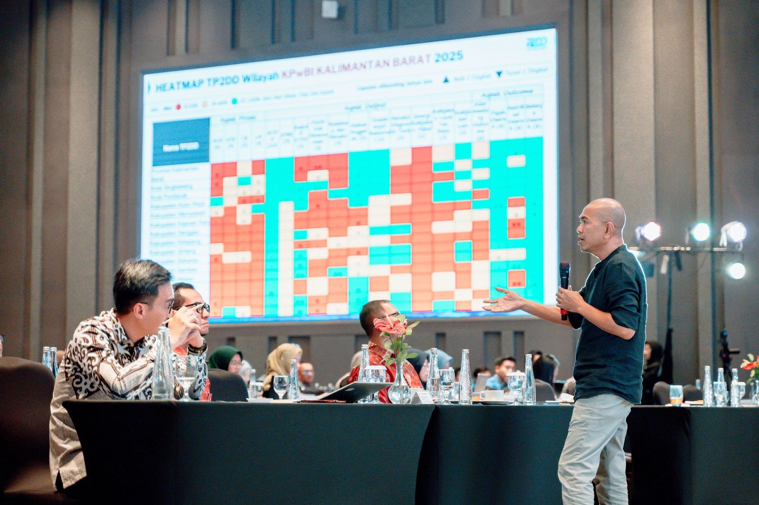 A presenter stands before a large heatmap data visualization screen while addressing seated conference attendees.