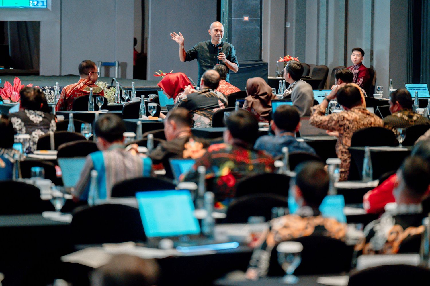 A speaker presents to a large audience at a professional conference, with attendees using laptops in a modern auditorium.