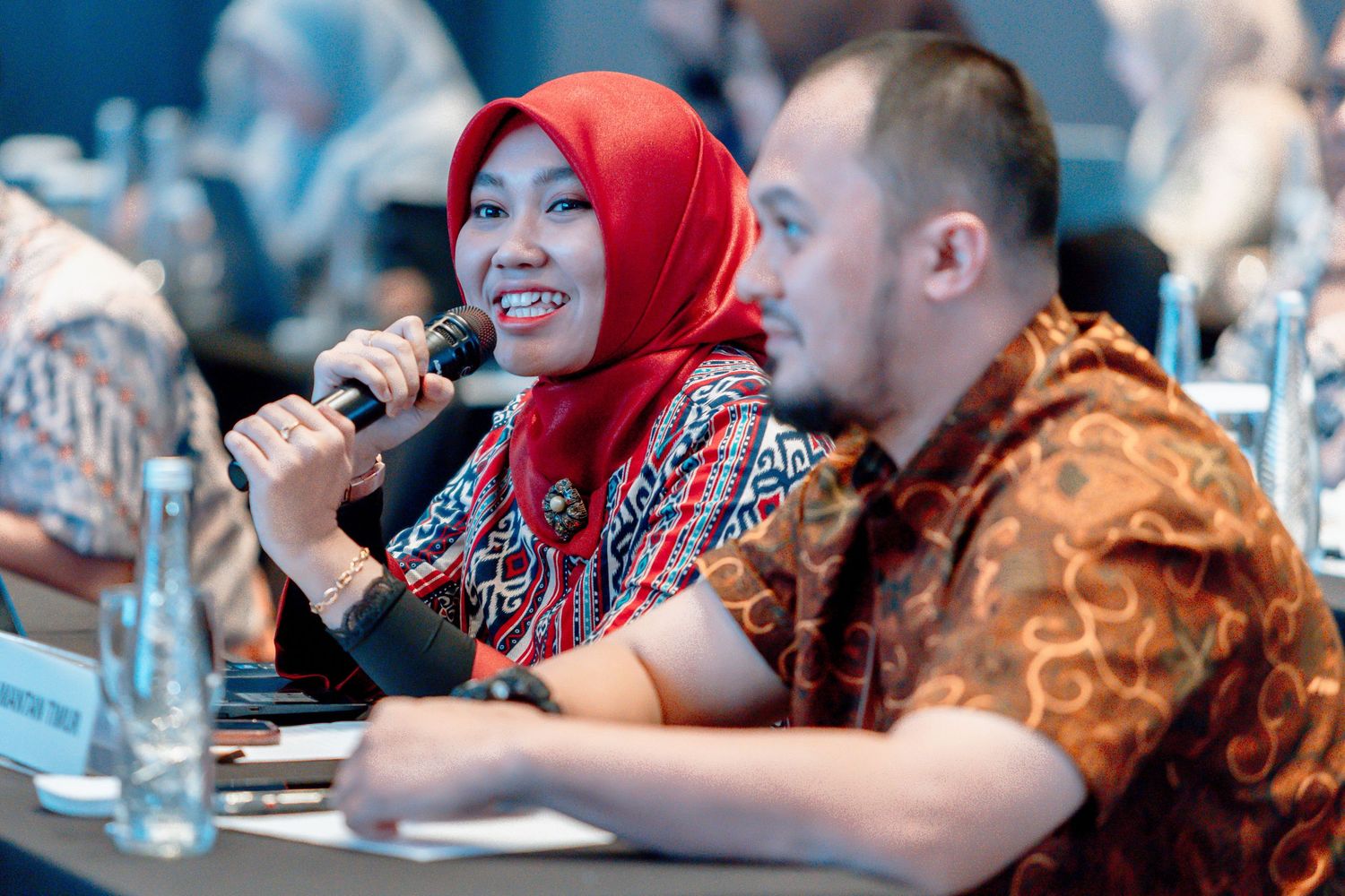 Woman in red hijab speaking into microphone at conference panel, seated beside man in batik shirt.
