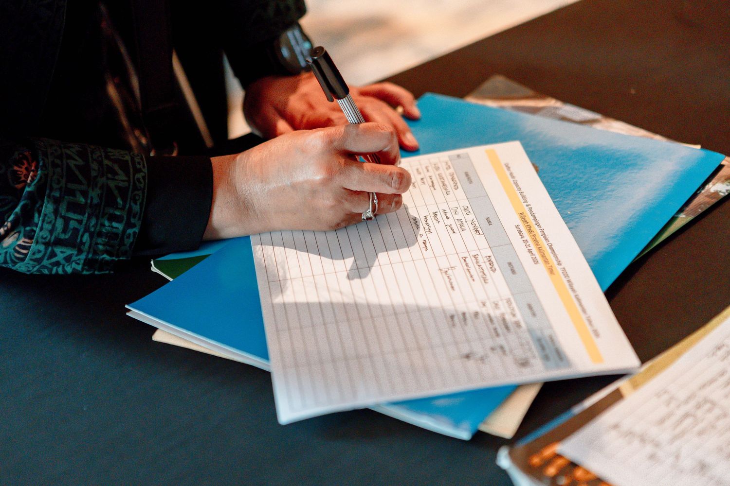 Person filling out a form with a pen on a stack of blue folders and documents on a table.