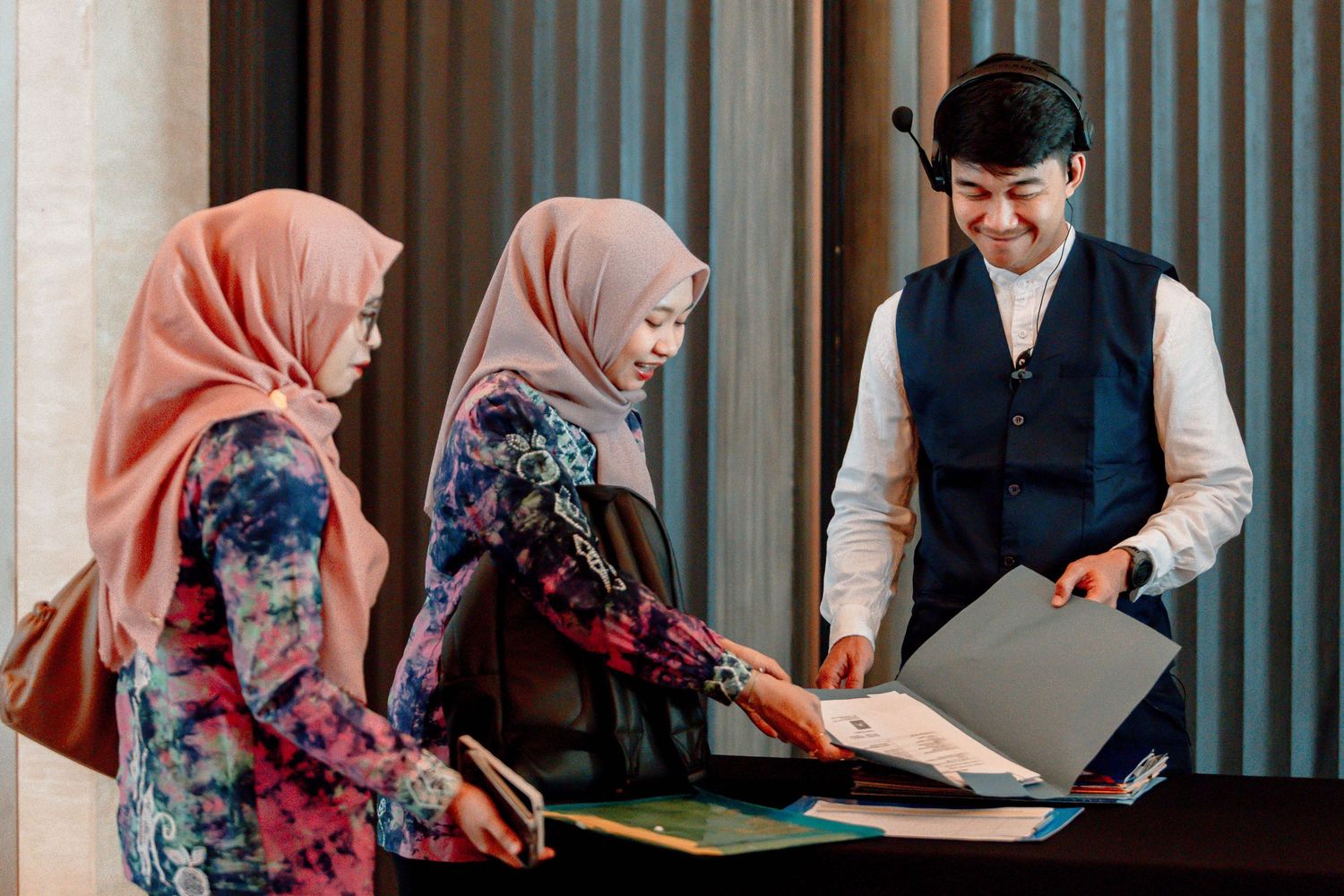 Hotel concierge smiling while assisting two women wearing hijabs at a reception desk with brochures.