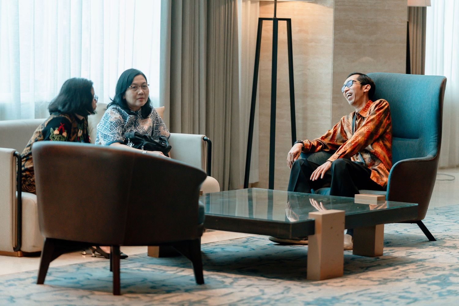 Three people in batik attire having a conversation in a modern hotel lobby lounge with elegant furniture.