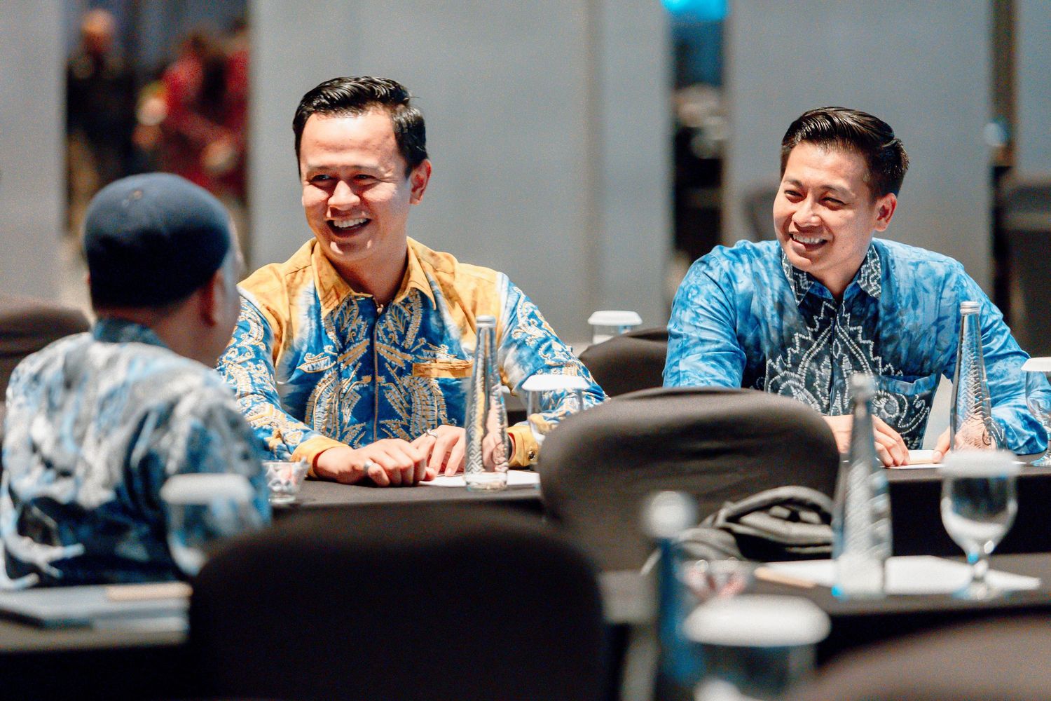 Three men in batik shirts laughing together at a business meeting table with glassware.