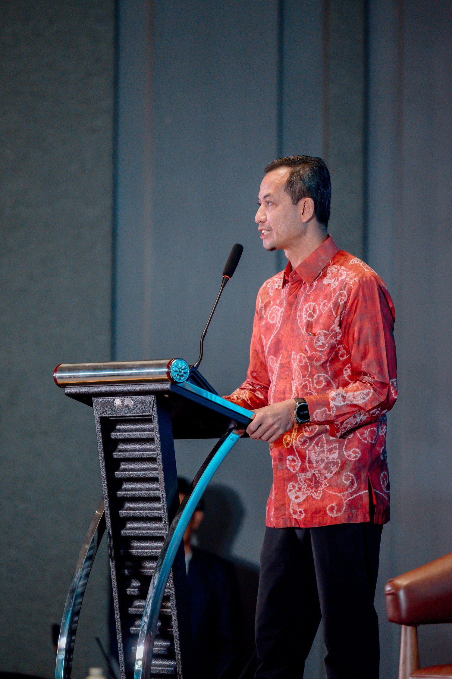 Man in red batik shirt speaking at podium with microphone during formal event or conference.