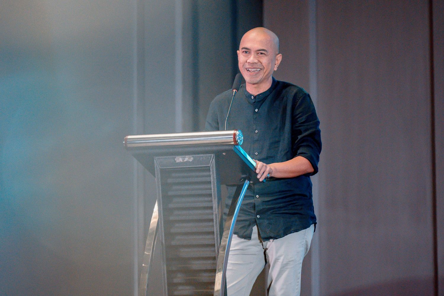 Man in black shirt smiling at podium during a professional speaking engagement or conference presentation.