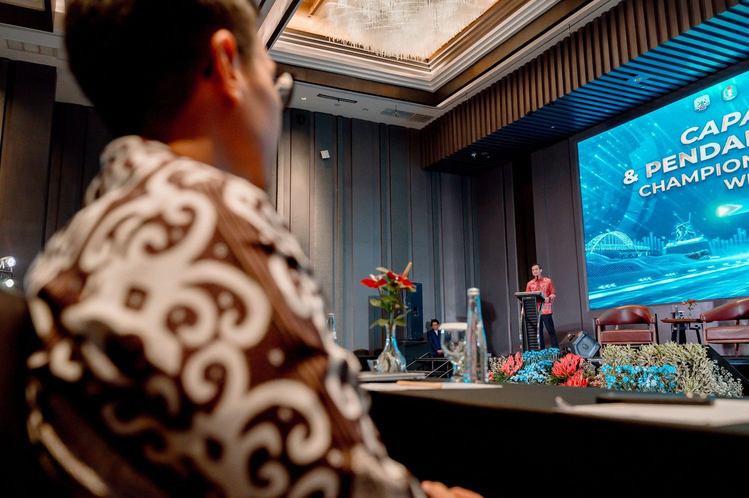 A speaker presents on stage at a formal conference with a large blue screen displaying championship event details.