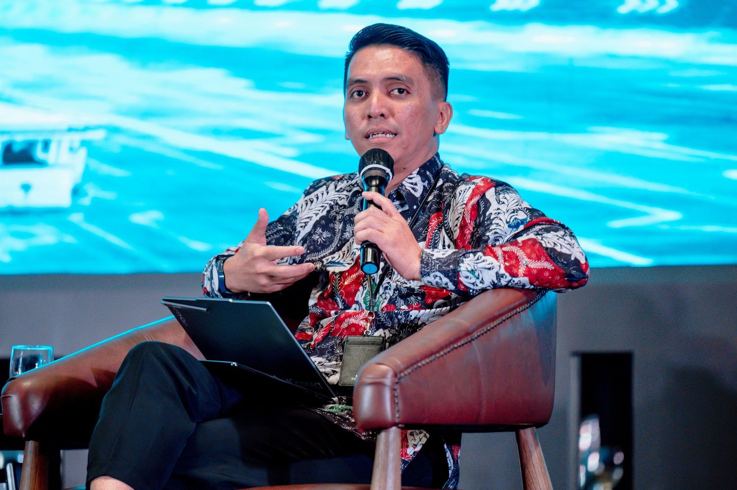 Man in batik shirt speaking at panel discussion, holding microphone and laptop at conference event.