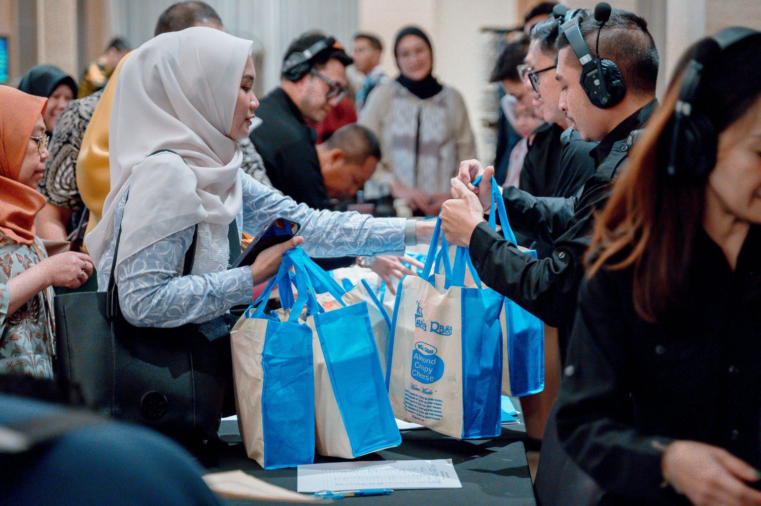 People exchanging blue branded tote bags at an indoor event or conference gathering.