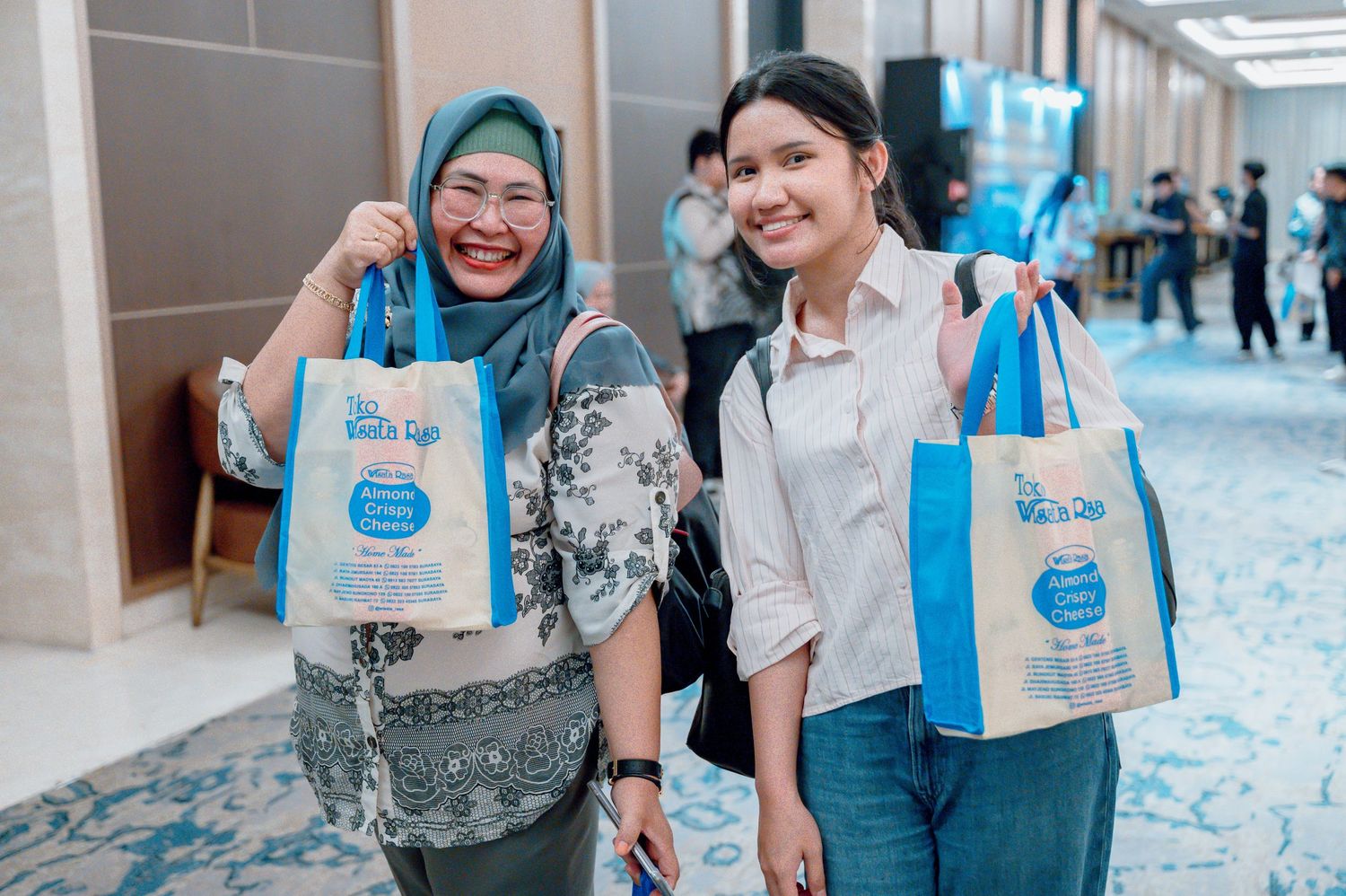 Two women smiling and holding blue branded tote bags at an indoor event in a modern venue.