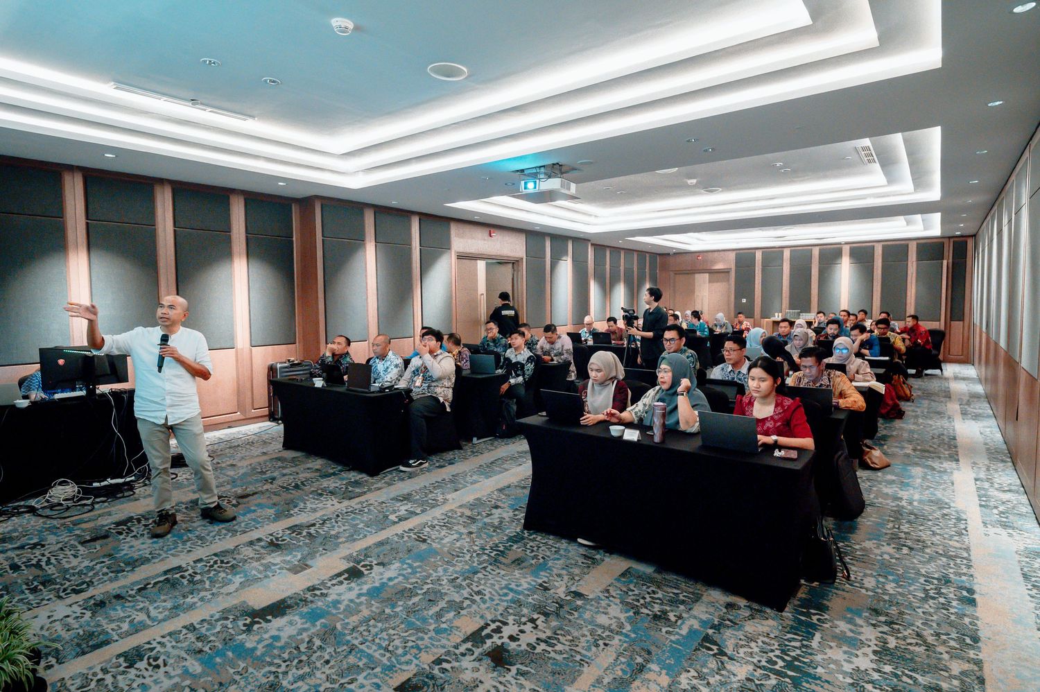 A presenter speaks to an audience in a modern conference room with blue patterned carpet.