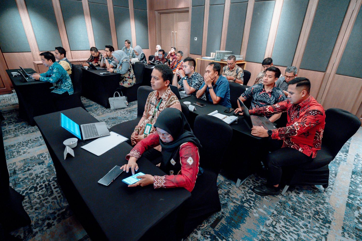 Participants in traditional batik attire attend a workshop session with laptops at black tables.