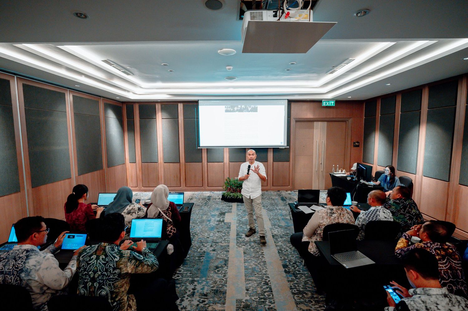 A trainer presents to attendees with laptops in a dimly lit seminar room with a projected screen.
