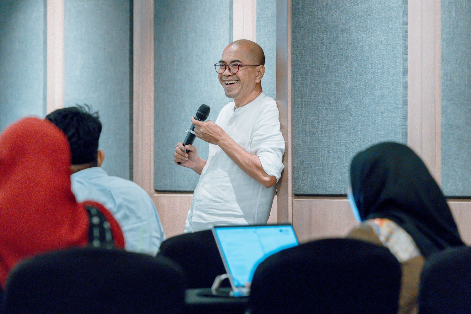 Bald man with glasses speaking into microphone while presenting to an audience in a modern classroom setting.