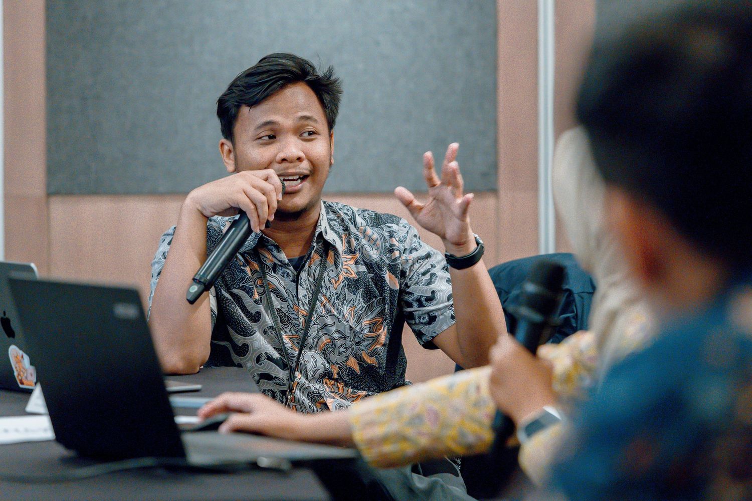 Man in batik shirt speaks into microphone at conference table with laptops, gesturing expressively during a meeting.