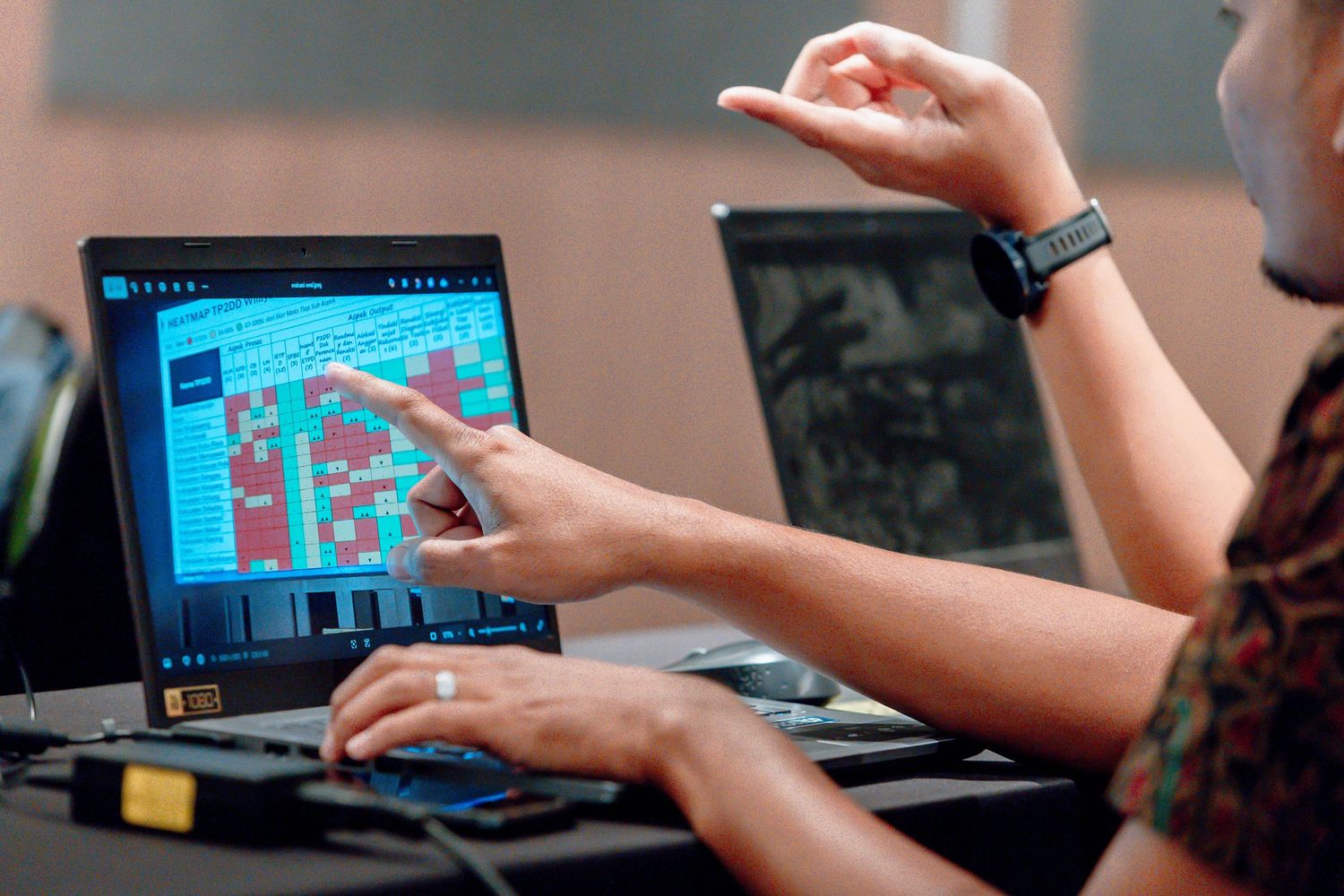 Two people collaborating on a laptop displaying colorful grid software, one pointing at the screen in a modern workspace.